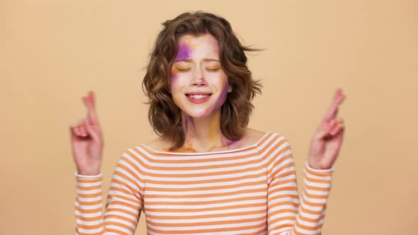 Portrait of Concentrated Woman with Multicolored Makeup Stains on Face Holding Fingers Crossed As