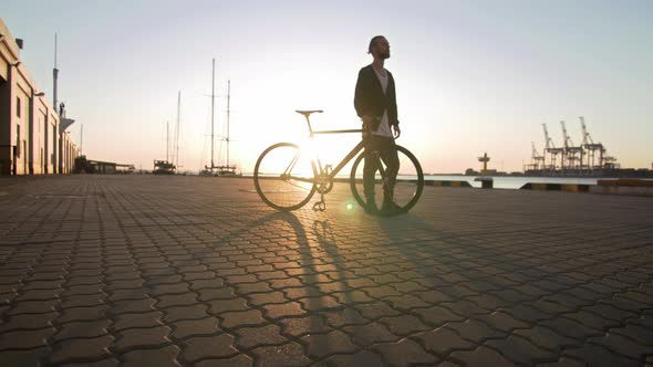 Young Hipster Man Walking with Bicycle During Sunset or Sunrise with Sea Port on Background