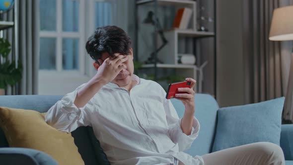 Asian Man Being Upset Losing Game On Smartphone While Sitting On Sofa In The Living Room