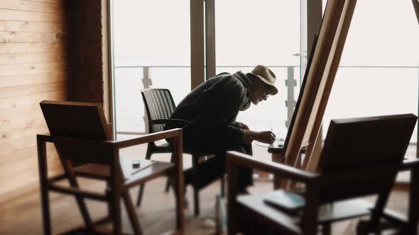 Middle Aged Artist Is Painting a Picture, Sitting in His Workshop