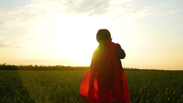 A Happy Child in a Superhero Costume Runs Across a Green Lawn Toward Sunset