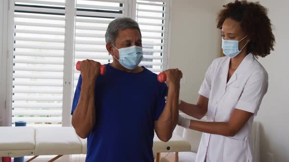 Mixed race female physiotherapist wearing mask helping senior exercise using dumbbells