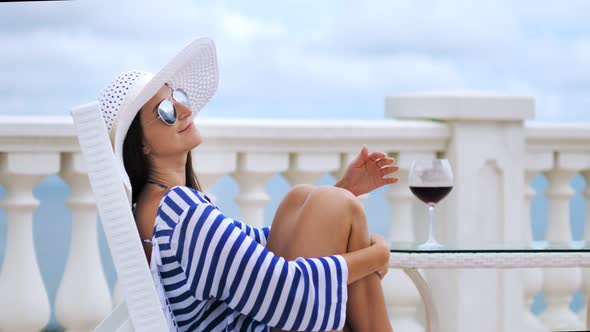 Elegant Woman in Sunglasses and Hat at Balcony Sitting on Chair Hugging Legs By Crossed Hands