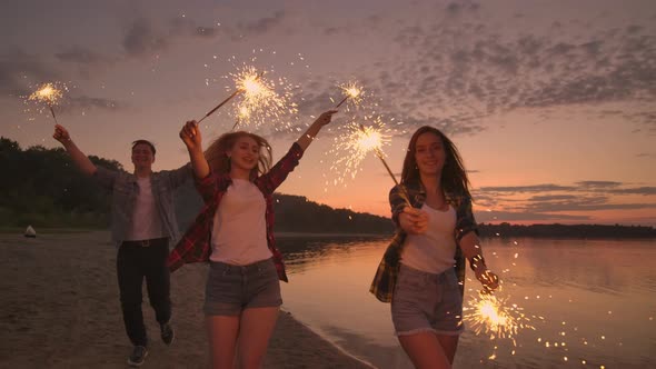 Cheerful Male and Female Friends are Running Along the Beach at Sunset Holding Sparkling Fireworks