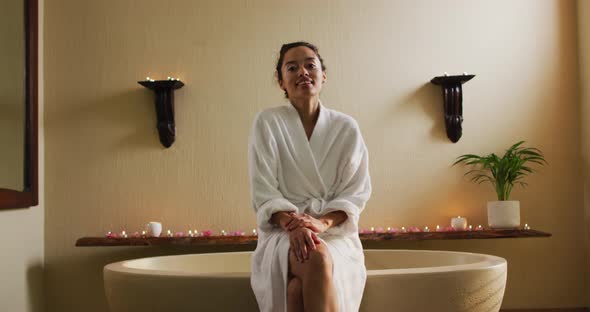 Happy biracial woman sitting in robe on bathtube