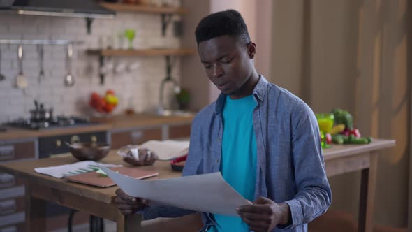 Focused Thoughtful African American Young Man Examining Business Graph in Kitchen Putting Away