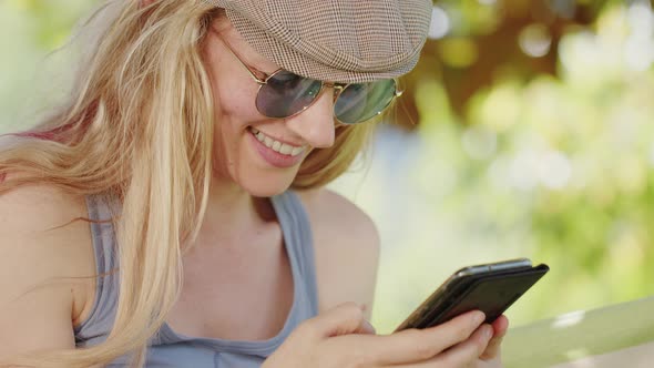 Smiling blonde woman with sunglasses using smartphone, lying relaxing on the hammock in the garden