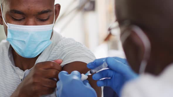 African american senior male doctor giving covid vaccine to male patient in home, wearing face masks