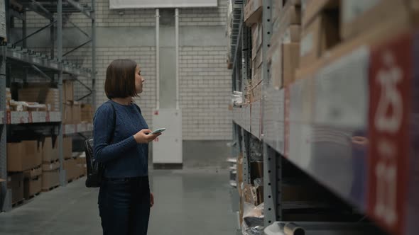 Young Caucasian Woman with Smartphone Choosing Goods in Shop Warehouse