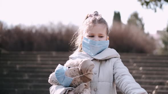 The Little Girl in a Disposable Mask Is Holding a Teddy Bear in a Mask Outside