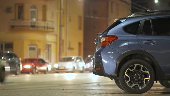 Blue car parked on brightly illuminated city street with moving traffic lights on background at