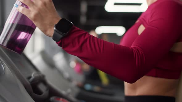 Woman Approaches Treadmill with Sports Bottle at Hand and Puts It in Cup Holder Closeup Side View