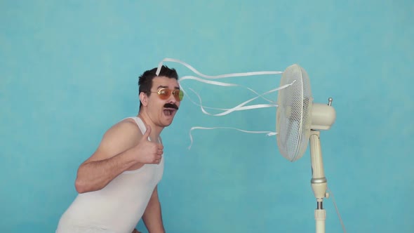 Funny Young Man with an Electric Fan, Enjoying the Cool Breeze Showing Thumbs Up on Blue Background