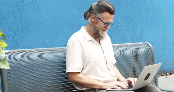 Middleaged Man Typing on a Laptop Sitting on a Bench in the Street