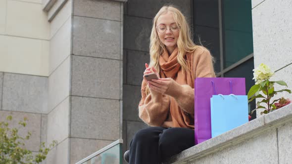 Female Shopper Sitting Outdoors Near Building with Colorful Bags Rejoices Discount Sale Black Friday