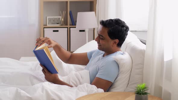 Happy Indian Man Reading Book in Bed at Home