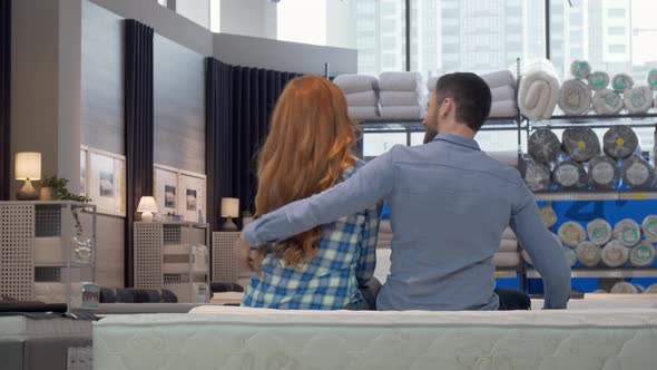 Rear View Shot of a Couple Hugging, Sitting on a New Bed at Furniture Store