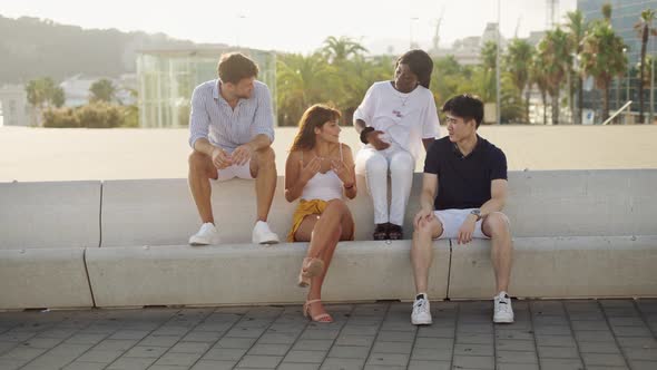 Four Young Multiethnic Friends Sitting on Concrete Bench Outdoors at Sunset and Communicating