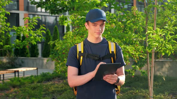 Zoom in Smiling Man Courier Food Delivery with Thermal Backpack Uses Tablet