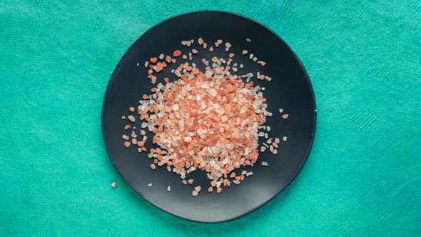 Crystals of pink Himalayan salt are falling into black ceramic plate