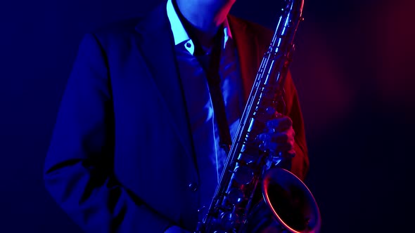 Male musician in suit and hat plays a saxophone in a studio under neon colored lighting. Saxophonist