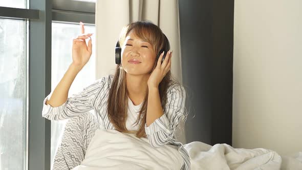 Happy Chinese Woman Listening To Music with Headphones Waking Up in the Morning in Bed From the Sun