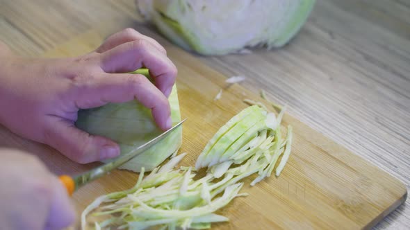 Woman is Finely Slicing Fresh Green Cabbage on Wooden Board with Sharp Knife in the Kitchen to