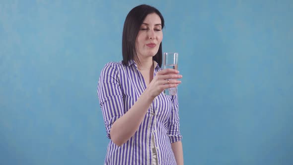 Young Woman with Pleasure Drinks Clean Filtered Water From a Glass Standing on a Blue Background