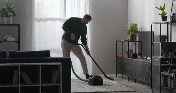 Happy Man Is Hoovering and Dancing Jokingly with Vacuum Cleaner in Living Room of Modern Apartment