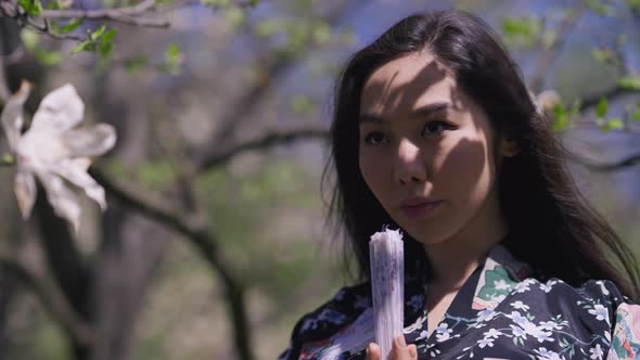 Portrait of Gorgeous Asian Young Woman Holding Closed Fan Looking Away in Sunlight Outdoors