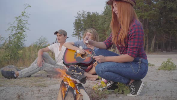 Young Hipster Friends Chilling on Beach Making Marshmallows with a Fire and Playing Guitar on the