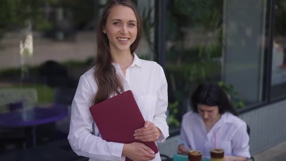 Portrait of Cute Confident Young Woman Posing Outdoors in Sidewalk Cafe with Blurred Friend Sitting