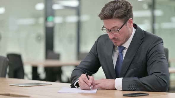 Focused Middle Aged Businessman Writing on Paper in Office 