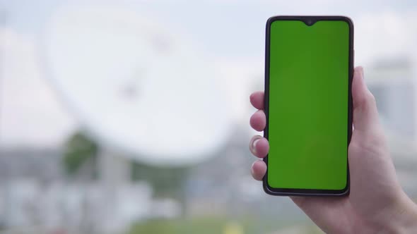 A Smartphone with Green Screen Is Held with a Satellite Dish in the Blurry Background - Closeup