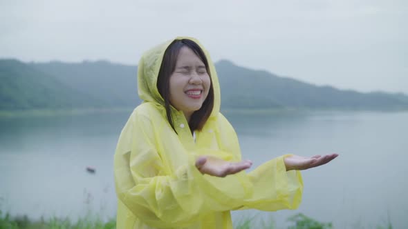 Young Asian woman feeling happy playing rain while wearing raincoat standing near lake.