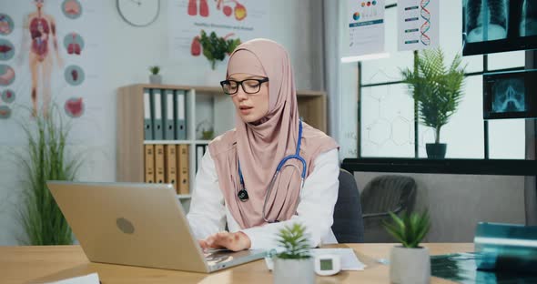 Female Doctor in Hijab Working on Computer in Her Workroom in Clinic and Looking Into Camera