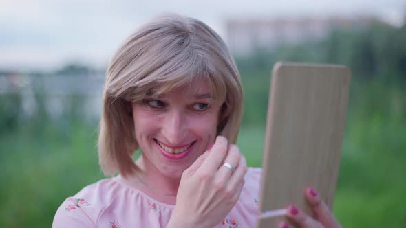 Cheerful Charming Trans Woman Adjusting Makeup Looking at Hand Mirror Standing Outdoors on Green