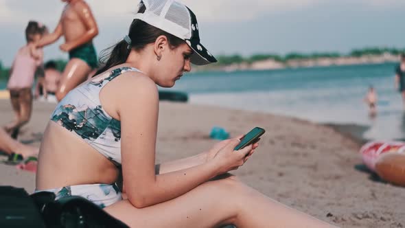 Beautiful Girl in a Bathing Suit Sits on a Sandy Beach with a Phone in Her Hands