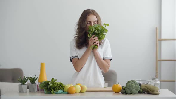 Young Smiling Woman Sniffing Fresh Greens Standing at the Table in Modern Kitchen. Concept of