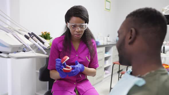 Pretty African Woman Dentist Talking to Her Male Patient Showing Artificial Jaw and Explaining Ways