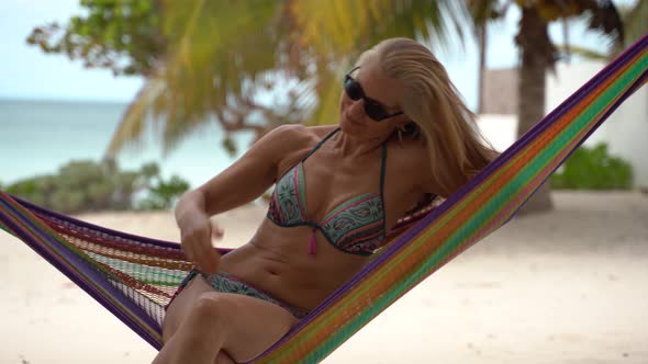Pretty mature woman in sunglasses and bikini in a hammock looking to the sky and then picking up her