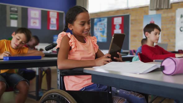 Mixed race schoolgirl in wheelchair, sitting in classroom using tablet, colleagues in background