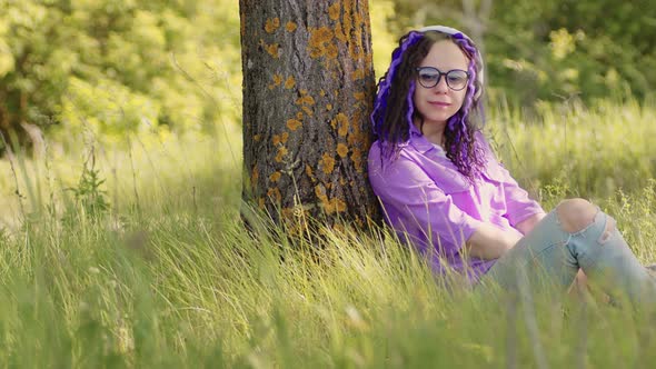 Young beautiful woman in glasses with wireless white headphones sitting on green grass near tree