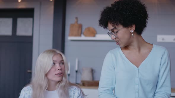 Young Girl Sits on a Table and Talks with Her Girlfriend Cutting Vegetables