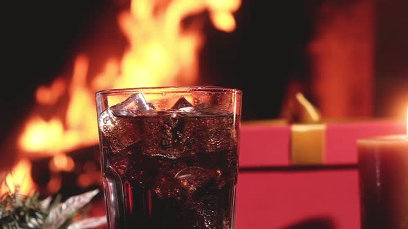 Carbonated Cola drink in glass with ice in front of burning fire in fireplace.