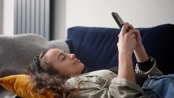 Young Smiling Woman Relaxing At Home Lying On Sofa Checking Social Media On Mobile Phone