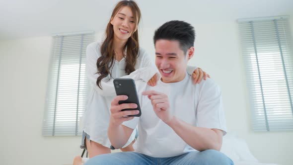 Asian young loving couple sit on bed and using mobile phone together.