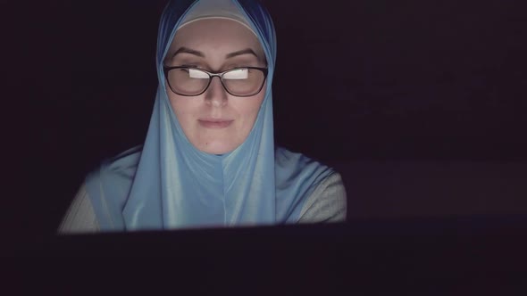 Portrait of Muslim Woman's Face in Hijab and Glasses at Night in the Dark Uses Computer