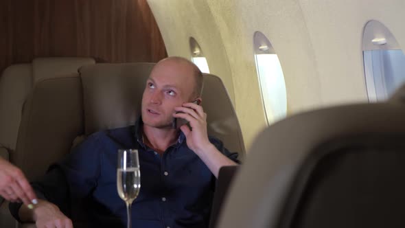A young Businessman Drinks Champagne sitting In his Business Jet, a Young Girl Brings him Champagne