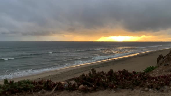 4k 60p, Golden sunset over ocean waves lapping onto a beach with a man walking his dog. Cargo ships